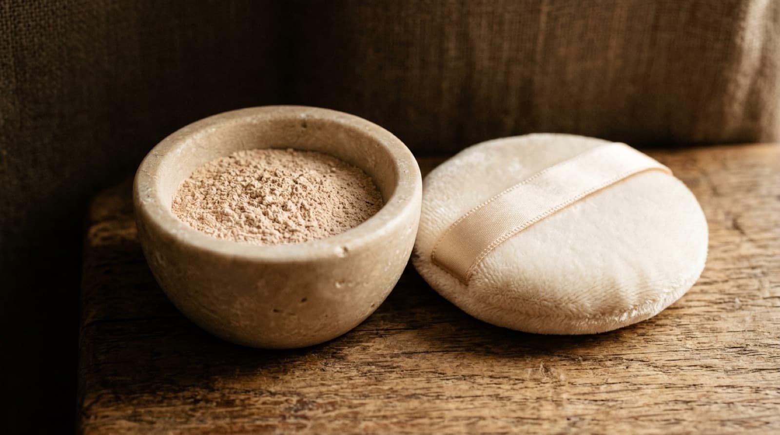 Poudre beige très fine dans une coupelle de pierre, à côté d'une houppette de velours.