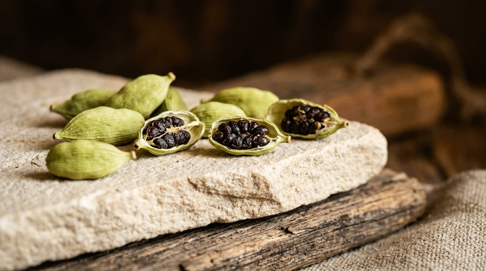 Capsules de cardamome verte, dont certaines ouvertes sur leurs graines noires, sur une pierre claire.