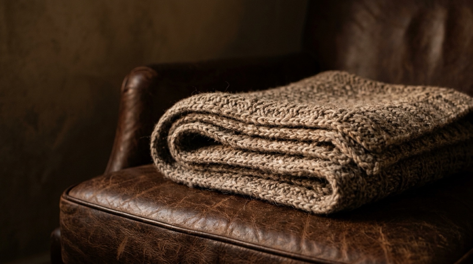 Écharpe de laine taupe pliée sur un fauteuil de cuir brun.